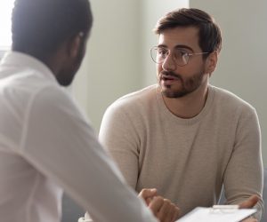 Man consulting with practitioner, African psychologist holding clipboard with card sitting in front of patient listens his mental health complaints. Job interview process applicant and HR manager concept